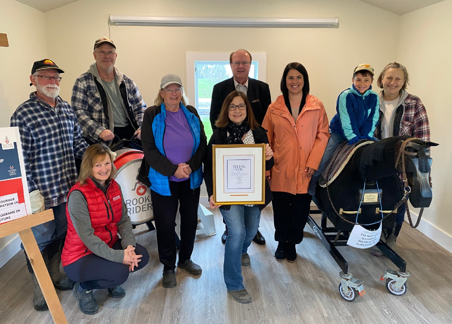 TEAD volunteers, staff, Board Members, and representatives of Donna Skelly smiling at the camera inside the clubhouse.  A Board Member is holding a thank you plaque for OTF. 