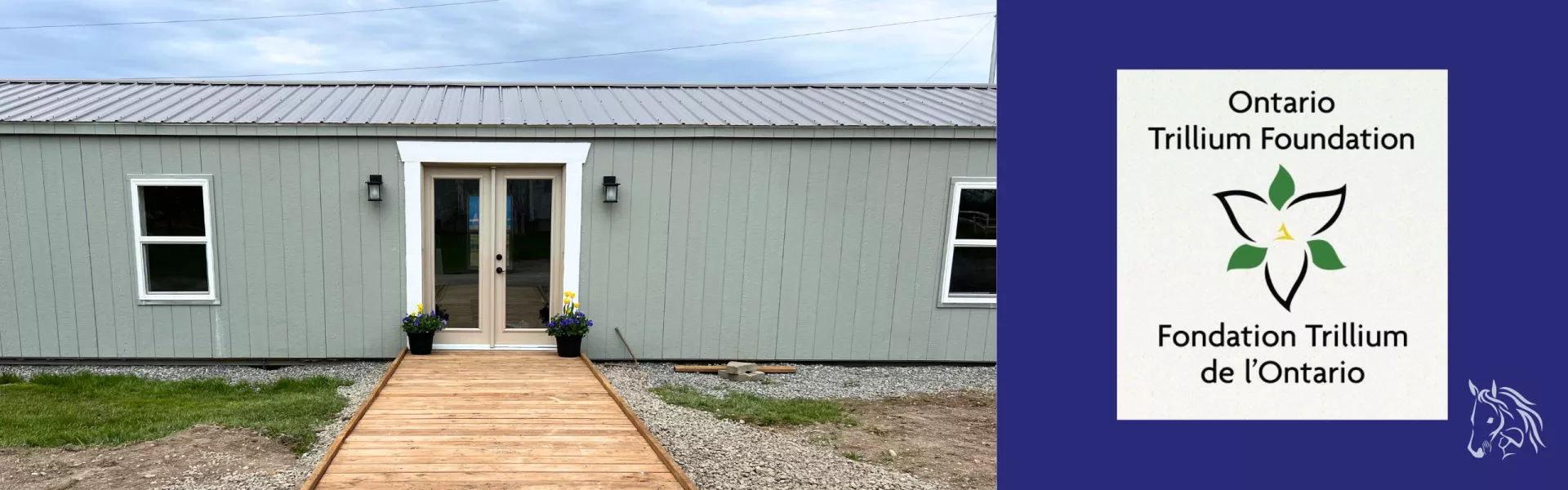 A blue-grey clubhouse with a wooden walkway leading to the entrance.  There are double doors with a white frame around them, and windows on either side.  And the Ontario Trillium Foundation logo: a white trillium flower with three green leaves on each side. 